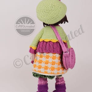 May include: A handmade crochet doll wearing a green beret, jacket, and a checkered skirt in orange and white. The doll has a pink purse and purple boots. The doll's design is by CAROcreated DESIGN.