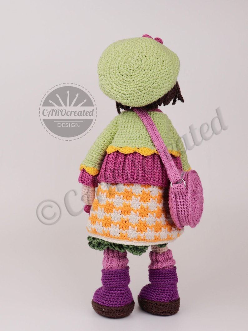 May include: A handmade crochet doll wearing a green beret, jacket, and a checkered skirt in orange and white. The doll has a pink purse and purple boots. The doll's design is by CAROcreated DESIGN.