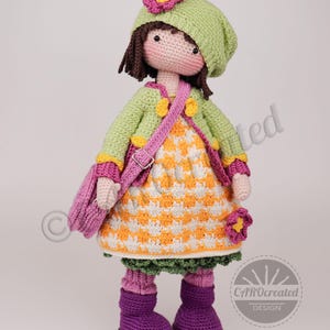 May include: A handmade crochet doll wearing a green jacket, hat, and boots. The doll has a pink purse and a dress with an orange and white pattern. The doll is approximately 12 inches tall.
