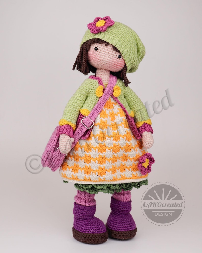 May include: A handmade crochet doll wearing a green jacket, hat, and boots. The doll has a pink purse and a dress with an orange and white pattern. The doll is approximately 12 inches tall.
