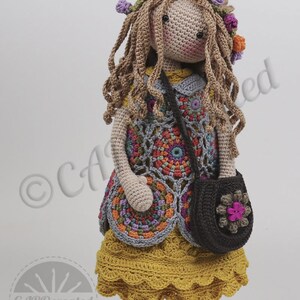 May include: Crocheted doll with long brown hair, wearing a colorful floral vest, a yellow skirt, and brown shoes. The doll is holding a brown bag with a pink flower.