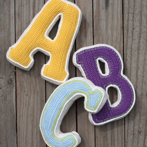 May include: Three crocheted letters, A, B, and C, are arranged on a wooden surface. The letter A is yellow, the letter B is purple, and the letter C is light blue.