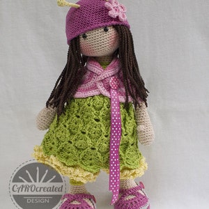 May include: A crocheted doll wearing a pink hat with a flower, a green dress with a pink ribbon, and pink shoes. The doll has brown hair and a pink and white cardigan.