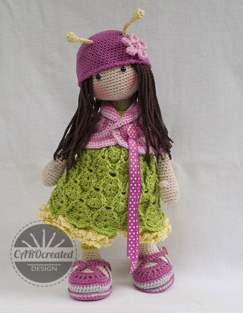 May include: A crocheted doll wearing a pink hat with a flower, a green dress with a pink ribbon, and pink shoes. The doll has brown hair and a pink and white cardigan.