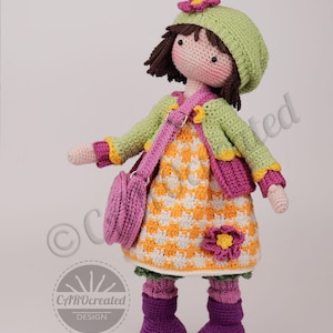May include: A handmade crochet doll wearing a green hat with a pink flower, a green jacket, and a yellow and white dress. The doll has a pink purse, purple boots, and brown hair. The doll is approximately 12 inches tall.