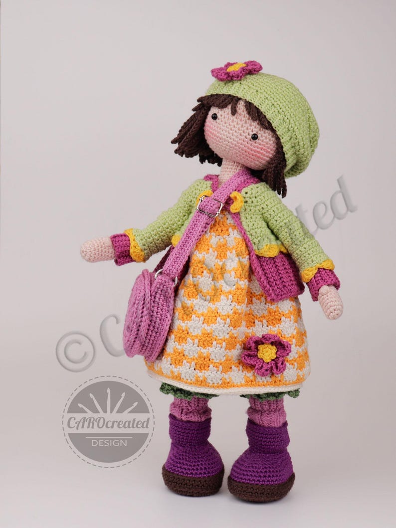 May include: A handmade crochet doll wearing a green hat with a pink flower, a green jacket, and a yellow and white dress. The doll has a pink purse, purple boots, and brown hair. The doll is approximately 12 inches tall.