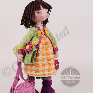 May include: A handmade crochet doll with brown hair, wearing a green jacket, orange and white dress, and purple boots. The doll has a matching purple purse. The doll is adorned with flower accents and is a charming, handcrafted item.
