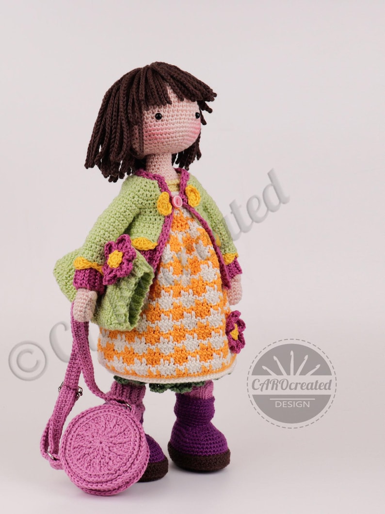 May include: A handmade crochet doll with brown hair, wearing a green jacket, orange and white dress, and purple boots. The doll has a matching purple purse. The doll is adorned with flower accents and is a charming, handcrafted item.