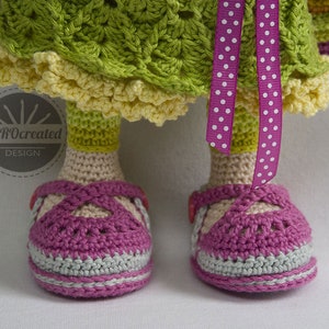 May include: Crocheted doll wearing a green and yellow dress with a pink polka dot ribbon. The doll is wearing pink and grey shoes.