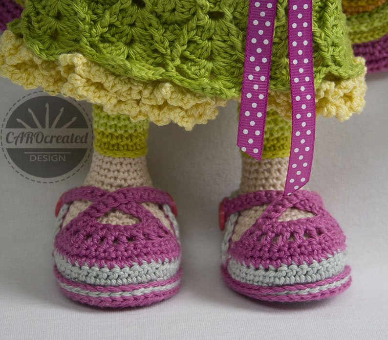 May include: Crocheted doll wearing a green and yellow dress with a pink polka dot ribbon. The doll is wearing pink and grey shoes.