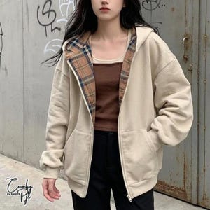 以下が含まれることがあります： ブラウン、ブルー、ホワイトのチェック柄の裏地が付いたフード付きのベージュのジップアップパーカー。長袖で、フロントジッパーと2つのポケットが付いています。ブラウンのタンクトップとブラックのパンツの上に着用されています。