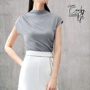 May include: A gray, short-sleeved top with a mock neck and a white skirt. The top has a draped design. The skirt has a silver belt. The text "Comfy by DANICA" is in the upper right corner.