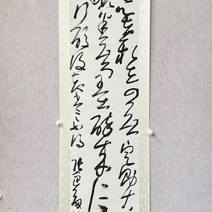 May include: A traditional Japanese calligraphy scroll with black ink on white paper. The scroll features a vertical arrangement of characters in a flowing, expressive style. The scroll is mounted on a light beige background with a decorative pattern.