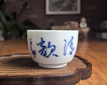 Pode incluir: Uma pequena chávena de chá de cerâmica branca com caligrafia chinesa azul. A chávena está sobre uma bandeja de madeira escura com uma superfície tecida. O fundo inclui uma mesa de madeira e uma pintura desfocada.