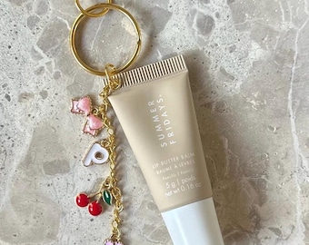 Könnte beinhalten: Eine beige Lippenbutter-Balsam-Tube mit dem Text "SUMMER FRIDAYS" und "LIP BUTTER BAIM" ist an einem goldenen Schlüsselanhänger befestigt. Der Schlüsselanhänger hat einen goldenen Verschluss, einen goldenen Ring und einen Charm mit rosa Schleifen, dem Buchstaben "P", Kirschen und einem rosa Herz.