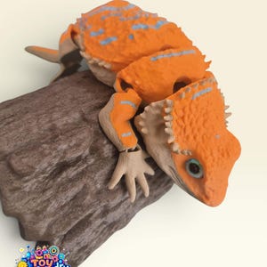 Peut inclure: Une figurine de lézard articulée orange et beige avec une surface texturée et bosselée, ornée de rayures bleu clair sur le dos et les membres. Le lézard est posé sur une base brune texturée en forme de bûche. Le logo Toy Foundry est visible.