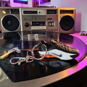 Può includere: Un portachiavi a forma di sneaker in miniatura con tomaia nera, intersuola bianca e dettagli arancioni, fissato a un anello metallico argentato e a un moschettone. Il design della sneaker include un logo swoosh nero e lacci testurizzati.