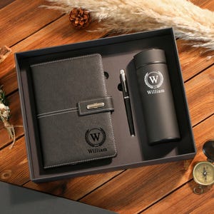 Peut inclure: Un coffret cadeau noir contenant un carnet, un stylo et un gobelet. Le carnet et le gobelet sont personnalisés avec la lettre "W" dans une couronne de laurier et le nom "William". L'ensemble est présenté dans une boîte noire sur une surface en bois.