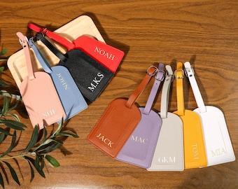 May include: Eight leather luggage tags with different names printed on them in white lettering. The tags are in various colors, including red, black, brown, purple, gray, yellow, and white.