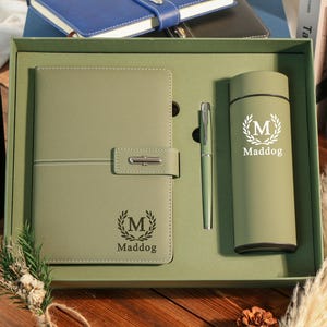 Peut inclure: Un coffret cadeau vert comprenant un carnet, un stylo et un gobelet. Le carnet a une fermeture à boucle et le texte "Maddog" avec un motif de couronne de laurier. Le gobelet porte également le texte "Maddog" et la couronne. Le stylo est argenté.