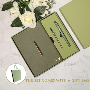Könnte beinhalten: Olivgrünes Notizbuch- und Stift-Set in einer Geschenkbox. Das Notizbuch hat einen goldfarbenen Metallakzent und Initialen mit einem Herz. Ein passender Stift und eine Mine sind enthalten. Das Set wird mit einer Geschenktüte geliefert.