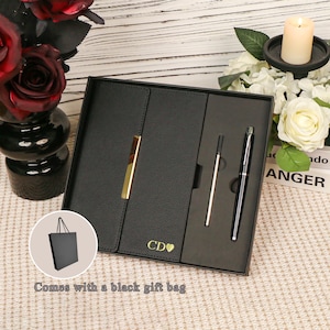 May include: A black gift set containing a notebook, a pen, and a stylus. The notebook features a gold clasp and the initials "CD" with a heart. Includes a black gift bag.