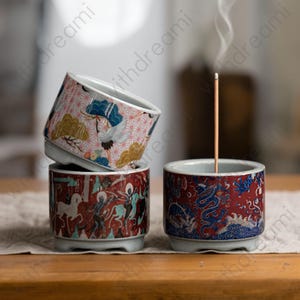 以下が含まれることがあります： 装飾的なセラミック香立てが3つ。1つは鶴と雲のピンク色の背景、もう1つは人物の赤い背景、3つ目は青い龍の赤い背景です。香立ての1つにはお香が焚かれています。