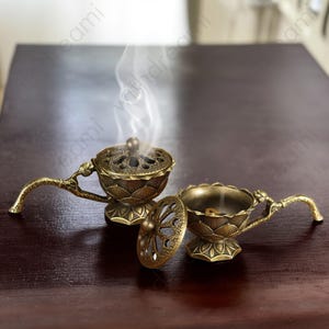 May include: Two antique brass incense burners with intricate lotus flower designs. One burner has its lid open, and smoke is rising from the incense. The burners have curved handles and sit on a dark wooden surface.