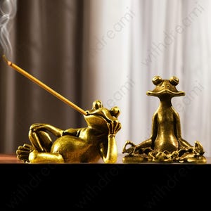 May include: Two brass frog figurines. One frog reclines, holding an incense stick, with smoke rising. The other frog sits in a meditative pose. Both are on a dark surface, with a blurred background.