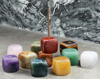 May include: A collection of cube-shaped gemstone incense holders in various colors, including red, green, yellow, and purple. A single incense stick is inserted into a red holder. The background features a marble wall.