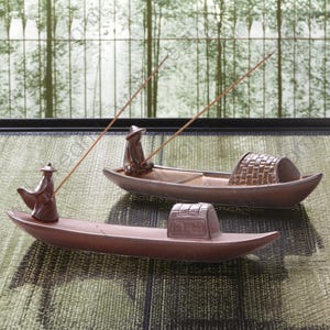 May include: Two brown ceramic incense burner boats with figures of people in them. The boats are on a woven mat. The incense sticks are sticking straight up.