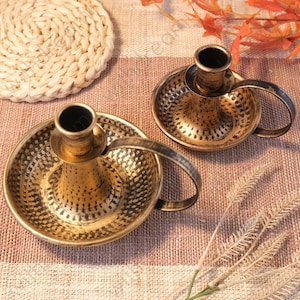May include: Two antique brass candle holders with handles. The candle holders have a hammered texture and a dark, aged finish. They sit on a striped, neutral-toned surface, with a woven coaster and dried foliage in the background. The candle holders are designed to hold a single candle.