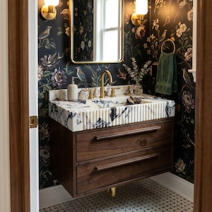 Può includere: Un mobile bagno sospeso in legno scuro con due cassetti e un piano in marmo bianco con venature nere. Il lavabo è integrato nel bordo scanalato in marmo. Sono presenti un rubinetto dorato e applique da parete coordinate.