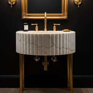 Könnte beinhalten: Ein weißes Marmor-Waschbecken mit goldenen Armaturen und Beinen. Das Waschbecken hat ein geriffeltes Design. Über dem Waschbecken hängt ein goldgerahmter Spiegel, und eine Seifenflasche steht auf der Theke.