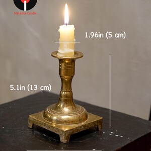 May include: A brass candlestick holder with a lit white candle. The holder has a square base measuring 3.54 inches wide and a total height of 5.1 inches. The candle has a diameter of 1.96 inches. The Natasha Candle logo is visible in the corner.