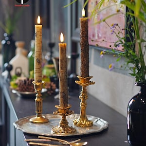 May include: Three lit candles with textured, bark-like exteriors sit in ornate gold candle holders on a silver tray. The candles are on a dark surface with gold utensils and a black vase with greenery.