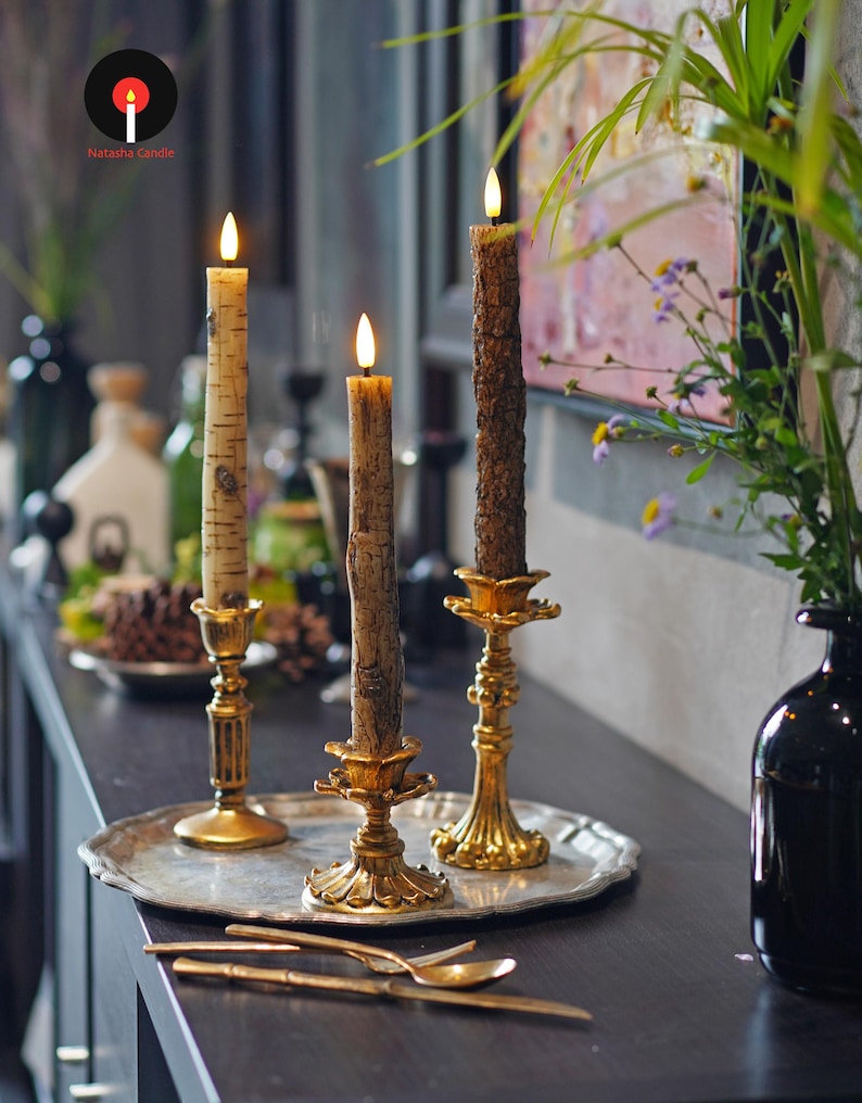 May include: Three lit candles with textured, bark-like exteriors sit in ornate gold candle holders on a silver tray. The candles are on a dark surface with gold utensils and a black vase with greenery.