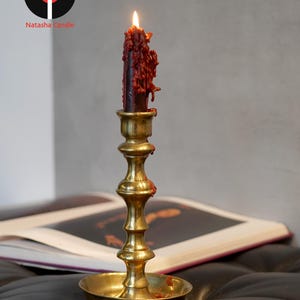 May include: A lit dark red candle with visible wax drips sits in a polished brass candlestick holder. The candlestick features a tiered, slender stem and a wide circular base. The logo for Natasha Candle is visible in the upper left corner.