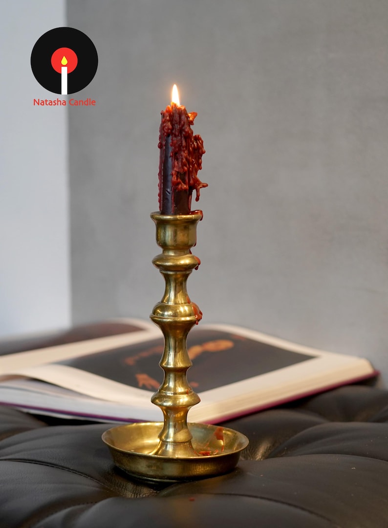 May include: A lit dark red candle with visible wax drips sits in a polished brass candlestick holder. The candlestick features a tiered, slender stem and a wide circular base. The logo for Natasha Candle is visible in the upper left corner.