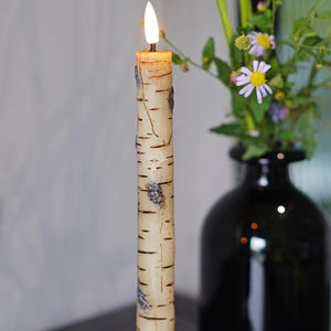 May include: A lit birch-style candle in a black candlestick holder. The candle has a realistic bark texture and is placed in front of a black vase with flowers. The candle is from Natasha Candle.