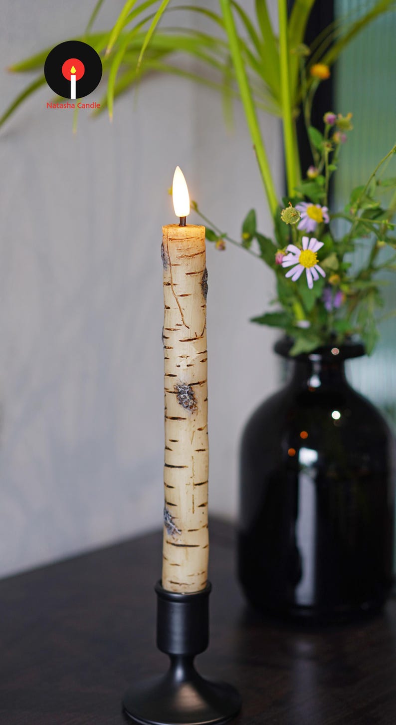 May include: A lit birch-style candle in a black candlestick holder. The candle has a realistic bark texture and is placed in front of a black vase with flowers. The candle is from Natasha Candle.