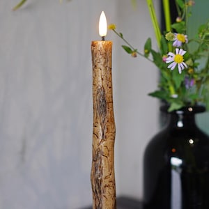 May include: A lit, textured candle with a bark-like appearance, set in a black candlestick. The candle has a warm, flickering flame. A black vase with flowers is in the background. The logo for Natasha Candle is in the upper left corner.