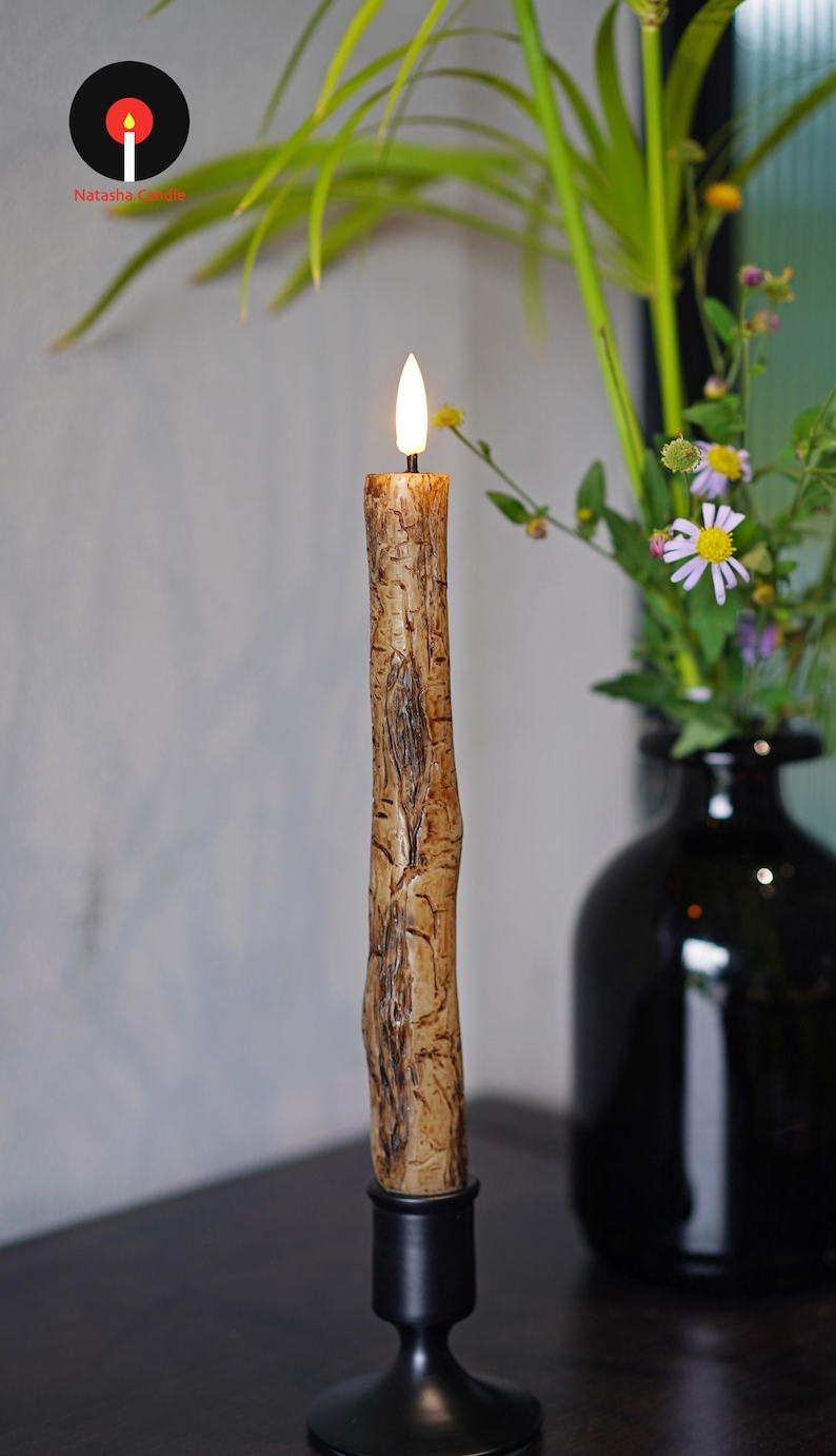 May include: A lit, textured candle with a bark-like appearance, set in a black candlestick. The candle has a warm, flickering flame. A black vase with flowers is in the background. The logo for Natasha Candle is in the upper left corner.