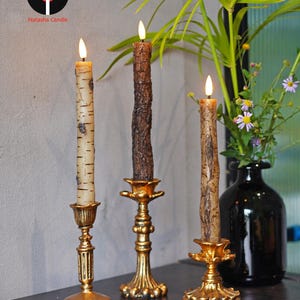 May include: Three decorative candles with flickering flames, each in a different style. The candles are set in ornate gold-colored candle holders. The candle on the left has a birch bark design, the middle one has a textured brown bark design, and the right one has a birch bark design. The logo "Natasha Candle" is in the upper left corner.