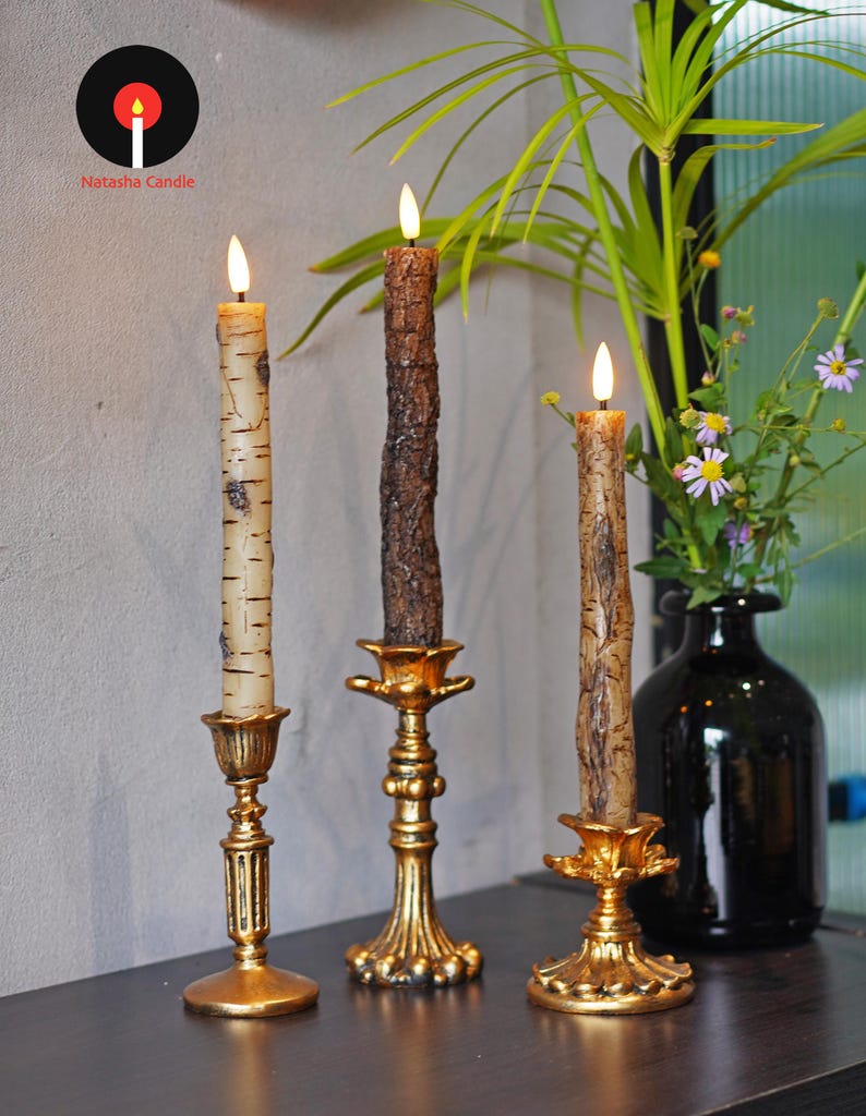 May include: Three decorative candles with flickering flames, each in a different style. The candles are set in ornate gold-colored candle holders. The candle on the left has a birch bark design, the middle one has a textured brown bark design, and the right one has a birch bark design. The logo "Natasha Candle" is in the upper left corner.