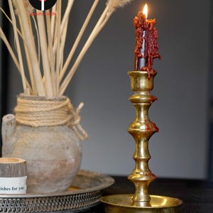 May include: A tall, polished brass candlestick holder with a tiered, cylindrical stem and a wide circular base. A dark red taper candle is lit, with thick, melted wax dripping down the sides of the candle and onto the brass stem and base.
