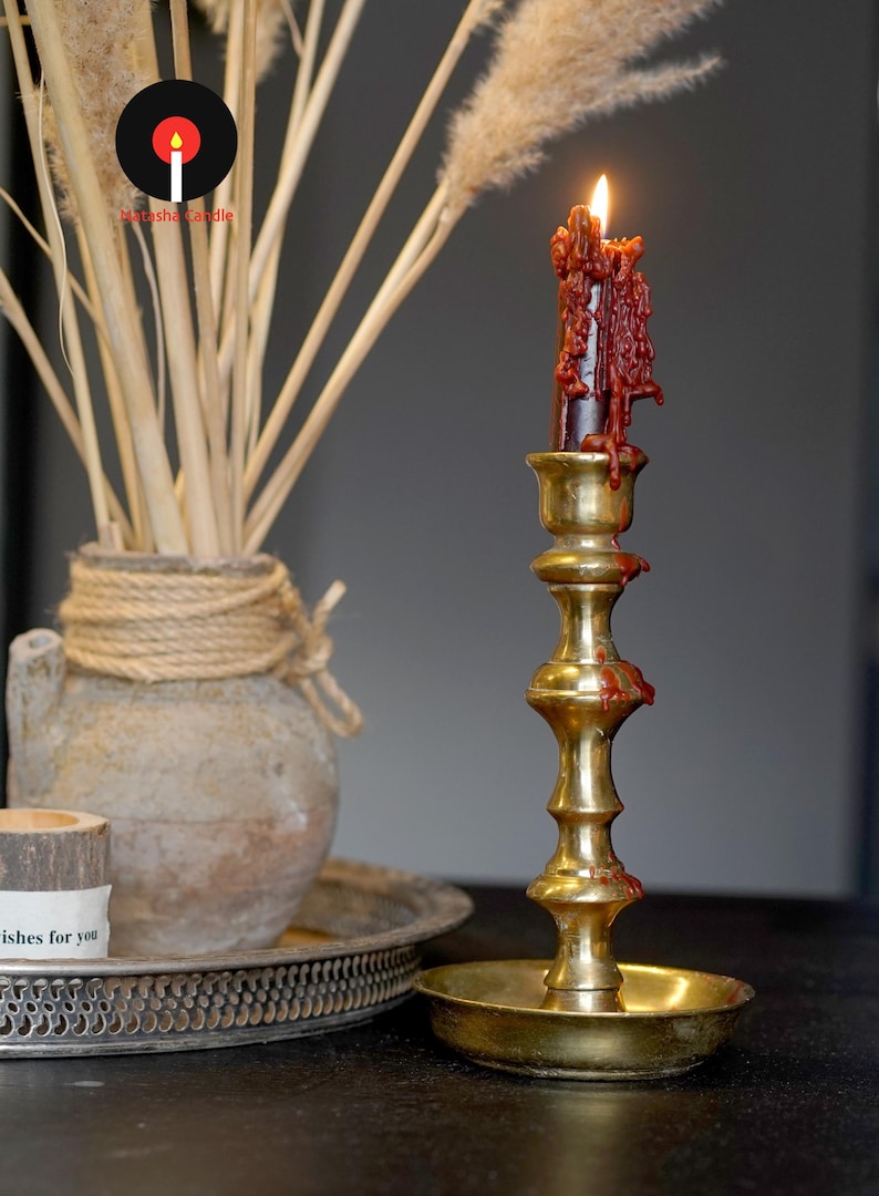 May include: A tall, polished brass candlestick holder with a tiered, cylindrical stem and a wide circular base. A dark red taper candle is lit, with thick, melted wax dripping down the sides of the candle and onto the brass stem and base.