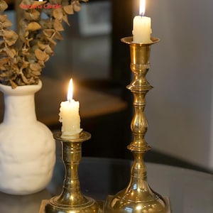 May include: Two polished brass candlesticks of varying heights, each featuring a square base and a slender, turned stem. Each candlestick holds a lit, off-white pillar candle with a visible flame. The metal surfaces reflect light.