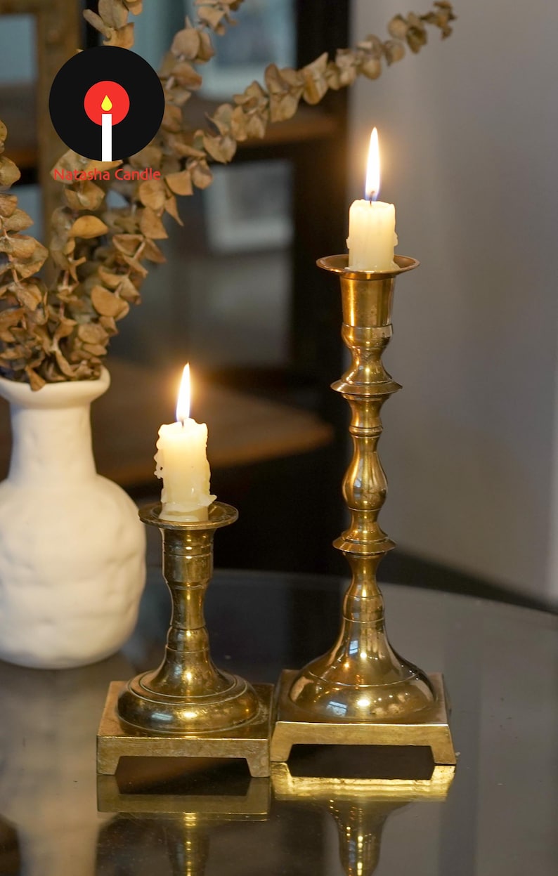 May include: Two polished brass candlesticks of varying heights, each featuring a square base and a slender, turned stem. Each candlestick holds a lit, off-white pillar candle with a visible flame. The metal surfaces reflect light.