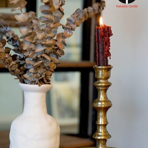 May include: A lit dark red taper candle with visible wax drips sits in a polished brass candlestick holder. Next to it is a white ceramic vase containing dried eucalyptus branches. The logo Natasha Candle is visible in the upper right corner.